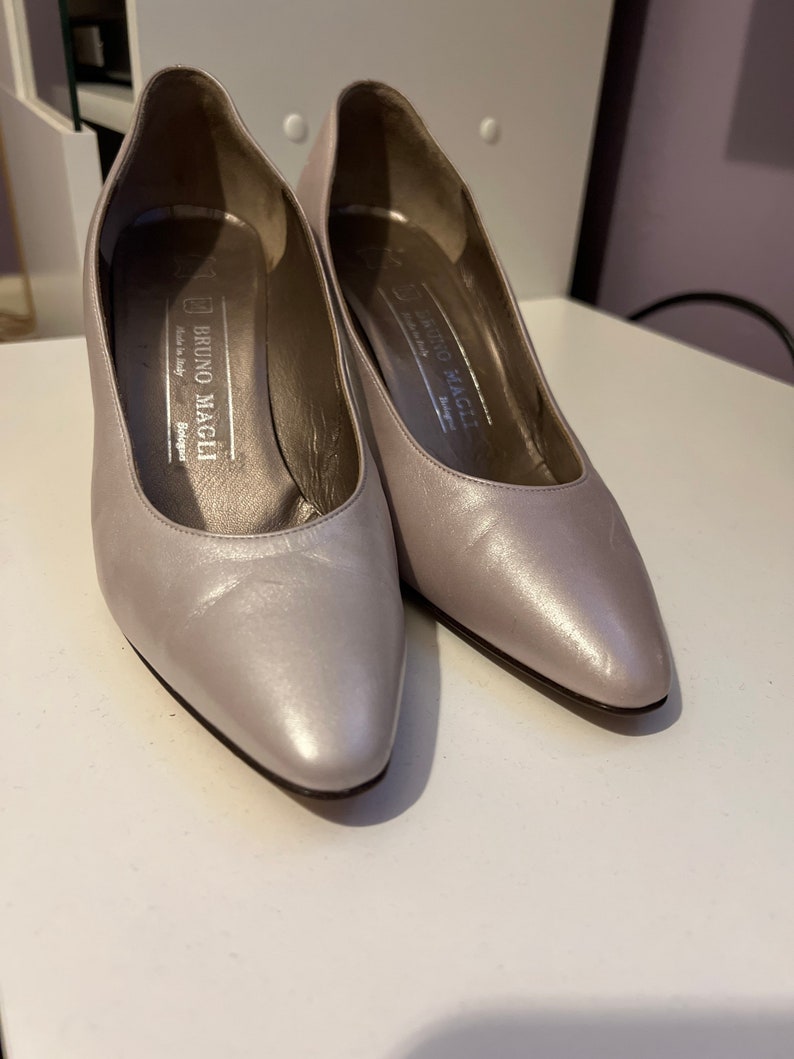 May include: A pair of silver leather pumps with pointed toes. The shoes have a small heel and are labeled "Bruno Magli" on the inside.