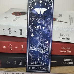 Puede incluir: Un marcapáginas rectangular con fondo azul oscuro estrellado que presenta dibujos de líneas blancas de una mariposa, un libro abierto con flores y dos manos extendidas hacia arriba. El texto SMUT está en una cinta y THE READER abajo.