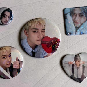 May include: A collection of five pinback buttons featuring images of a person. The buttons are round, square, and heart-shaped. The images have a variety of poses and expressions, with some holding a heart-shaped object.