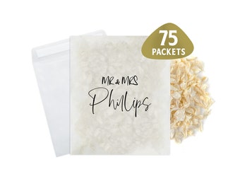 May include: A white paper bag with the text "MR & MRS Phillips" printed on it. The bag is filled with white confetti and is accompanied by a gold sticker that reads "75 PACKETS".
