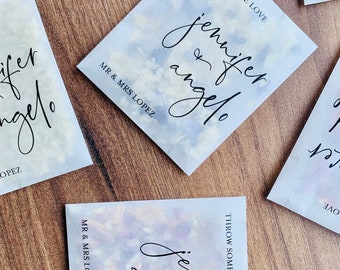 May include: A set of white paper favor bags with a watercolor design and the text "THROW SOME LOVE" and "MR & MRS LOPEZ" printed on them. The bags feature the names "jennifer & angelo" in black cursive writing.