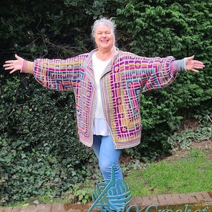 Könnte beinhalten: Eine Frau trägt eine bunte gehäkelte Strickjacke mit geometrischem Muster. Die Jacke hat eine Kapuze und ist vorne offen. Die Frau lächelt und hat die Arme ausgestreckt.