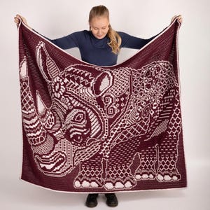 Pode incluir: Uma manta de crochê em tons de vinho e branco com um grande desenho de rinoceronte. A manta está sendo segurada por uma pessoa, mostrando os detalhes intrincados do design.