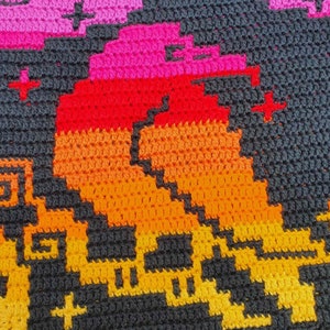 May include: A close-up of a crocheted blanket with a geometric pattern in shades of orange, red, pink, and black.
