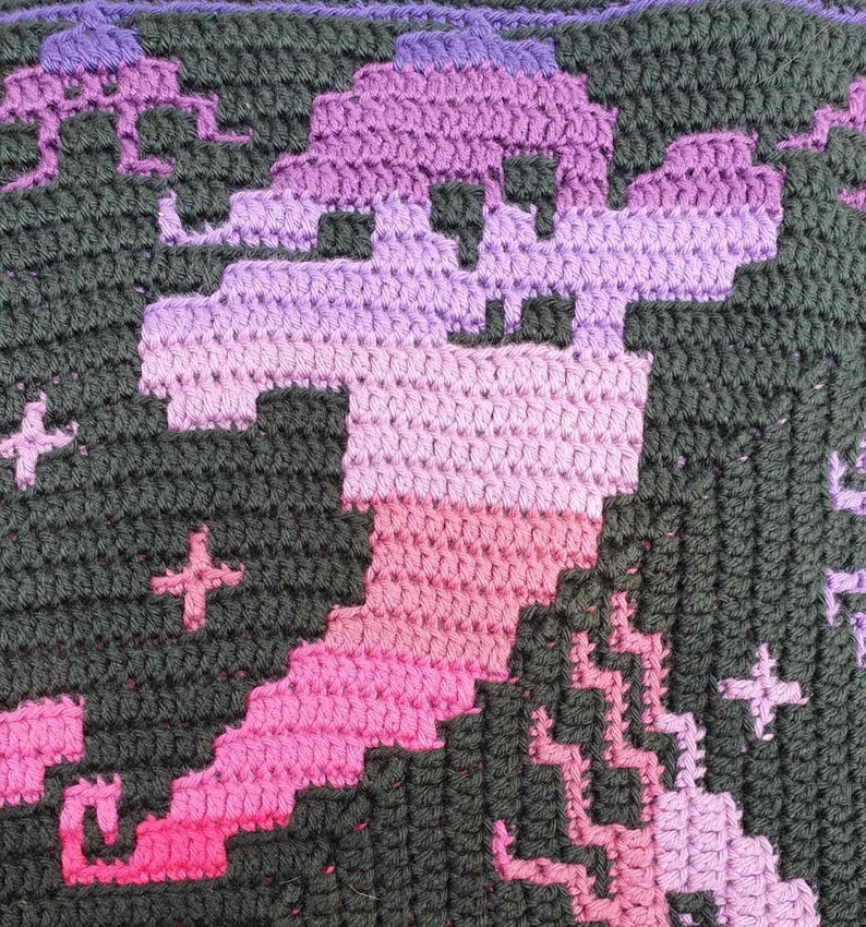 May include: A close-up of a crocheted blanket with a pixelated design of a dragon in shades of pink, purple, and black.