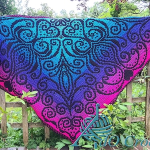 May include: A large, colorful crochet shawl with a black, swirling pattern. The shawl is made with a gradient yarn that transitions from pink to purple to blue to turquoise. The shawl is draped over a wooden fence.