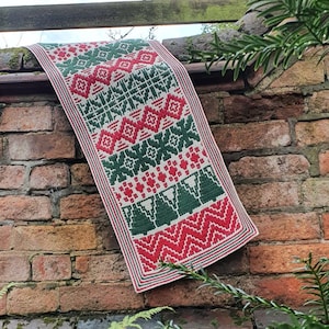 Op de afbeelding: Een rood, groen en witte gehaakte kerstboomloper met een geometrisch patroon. De loper hangt aan een bakstenen muur met groene planten op de voorgrond.