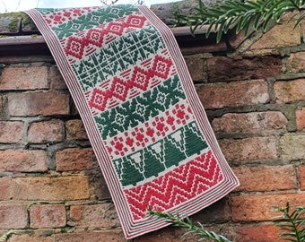 May include: A red, green, and white crocheted Christmas tree runner with a geometric pattern. The runner is hanging on a brick wall with green plants in the foreground.