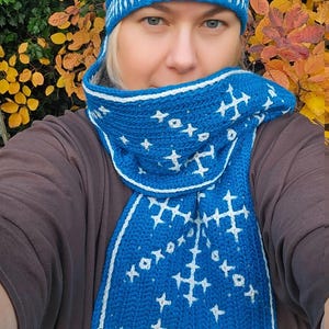 May include: A blue and white crochet hat with a white pom-pom and matching scarf. The scarf and hat feature a geometric pattern. The image also shows the text "Do Crochet".