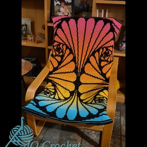 Könnte beinhalten: Eine farbenfrohe Häkeldecke mit Farbverlauf in Rosa, Orange, Gelb und Blau, mit schwarzen Details, drapiert über einem Holzstuhl. Die Decke hat ein florales Design und den Text "AdO Crochet".