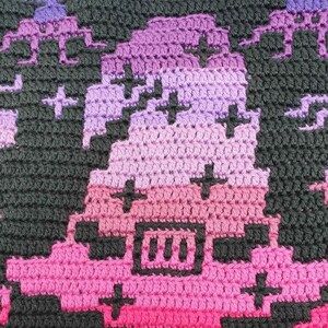 May include: A close-up of a crocheted piece with a gradient of pink, purple, and black. The piece features a pixelated design with a black square containing three vertical lines.