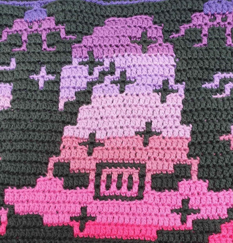 May include: A close-up of a crocheted piece with a gradient of pink, purple, and black. The piece features a pixelated design with a black square containing three vertical lines.