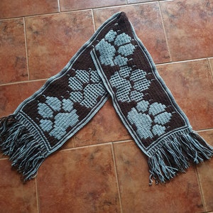 May include: A brown and blue crocheted scarf with a fringe on each end. The scarf features a repeating pattern of blue paw prints on a brown background.