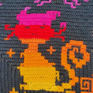May include: A crocheted wall hanging featuring a pixelated design of a cat in shades of orange, red, and pink against a dark gray background.