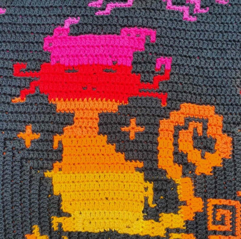 May include: A crocheted wall hanging featuring a pixelated design of a cat in shades of orange, red, and pink against a dark gray background.