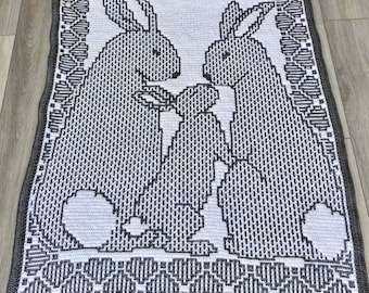 May include: A white and gray crochet blanket with a design of two bunnies. The blanket has a gray border with a repeating pattern of hearts. The text "Overlay mosaic crochet by Marissa Childs" is at the bottom of the blanket.