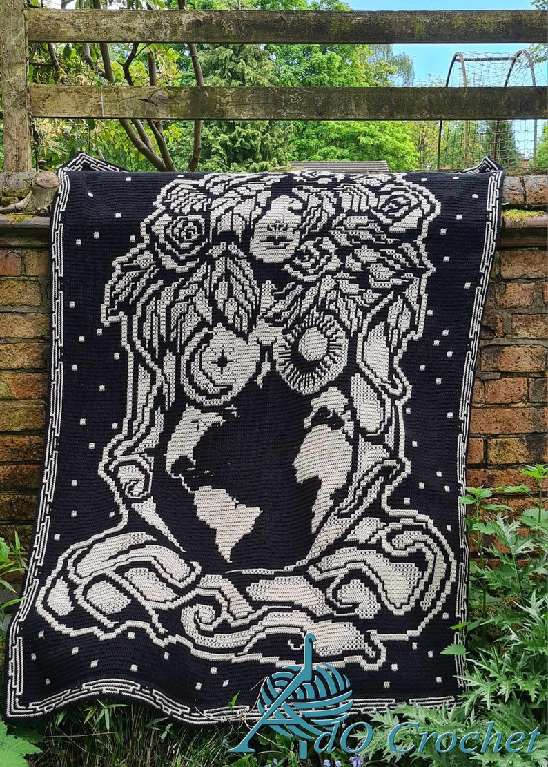 K&ouml;nnte beinhalten: Eine schwarze und wei&szlig;e H&auml;keldecke mit dem Gesicht einer Frau, umgeben von Blumen und Bl&auml;ttern. Die Frau h&auml;lt einen Globus in ihren H&auml;nden. Die Decke hat einen schwarzen Hintergrund mit wei&szlig;en Details.
