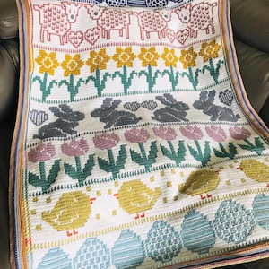 May include: A white crocheted blanket with an Easter theme. The blanket features a repeating pattern of lambs, daffodils, bunnies, tulips, chicks, and eggs. The blanket has a colourful border.
