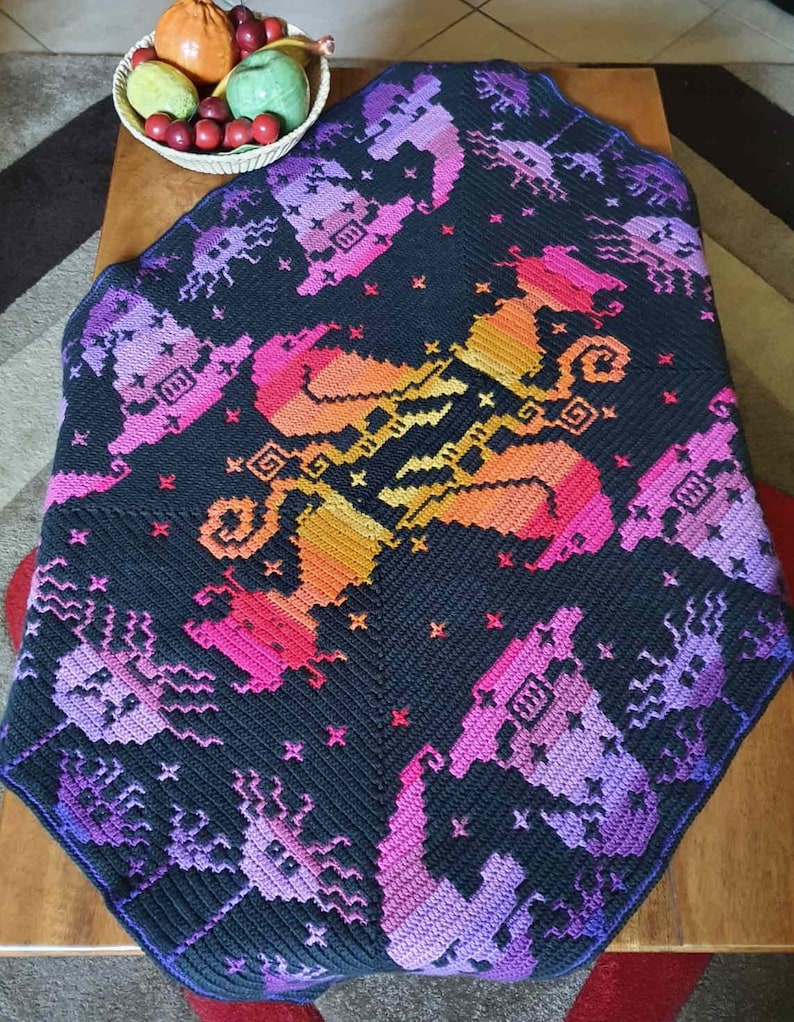 May include: A black crochet blanket with a colorful pixelated design of a unicorn. The blanket is made with a variety of shades of purple, pink, orange, and yellow yarn.