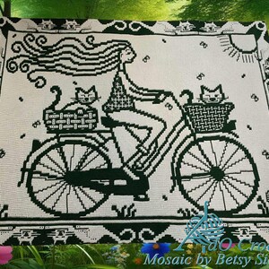 Puede incluir: Una manta de ganchillo blanca y verde con una mujer montando en bicicleta con dos gatos en cestas. El diseño incluye un sol, flores y un borde decorativo. La manta es un diseño de mosaico.
