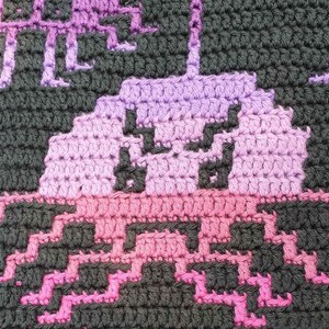 May include: Crocheted blanket with a pink and purple alien design. The alien is surrounded by stars.