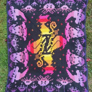 May include: A crocheted blanket with a colorful, pixelated design of a witch with a cauldron. The blanket is black with purple, pink, orange, and yellow accents. The border of the blanket features a repeating pattern of purple and black crocheted mushrooms.