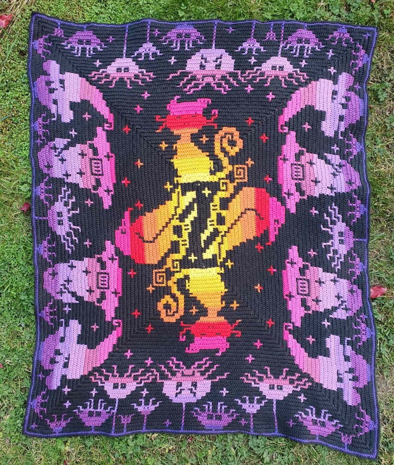 May include: A crocheted blanket with a colorful, pixelated design of a witch with a cauldron. The blanket is black with purple, pink, orange, and yellow accents. The border of the blanket features a repeating pattern of purple and black crocheted mushrooms.