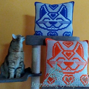 Pode incluir: Duas almofadas quadradas de crochê com rostos de gato, uma azul e outra laranja, estão em uma árvore para gatos. As almofadas têm fundos brancos e desenhos geométricos de gatos. Um gato malhado está sentado em uma prateleira inferior, olhando para cima.