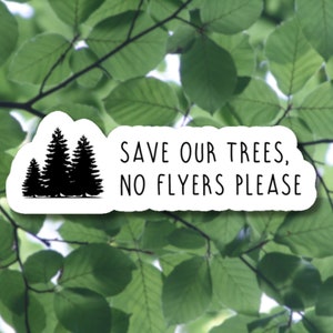 May include: A white sticker with a black silhouette of three pine trees and the text "Save our trees, no flyers please". The sticker is placed on a background of green leaves.