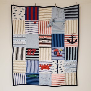 May include: A blue and white patchwork quilt made from baby clothes. The quilt features a variety of patterns, including stripes, polka dots, and nautical themes. The quilt is edged in a dark blue fabric.