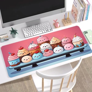 May include: A long, rectangular desk mousepad featuring a cartoon illustration of cupcakes and mochi. The cupcakes have various colors and toppings, including cherries. The background is pink, and the mousepad sits on a white desk.