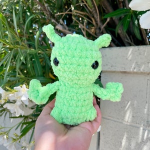 May include: A green crocheted alien plush toy with black eyes. The toy is being held in a person's hand.