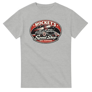 以下が含まれることがあります： ロケットとクラシックカーのグラフィックが特徴のライトグレーのTシャツ。赤と黒の楕円のデザインで「Rocket's Speed Shop, SO14, Hampshire」の文字が入っています。