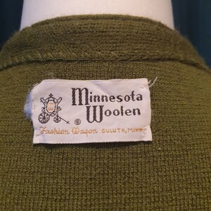 Op de afbeelding: Een groene wollen trui met een wit label waarop staat "Minnesota Woolen Fashion Wagon Duluth, Minn."