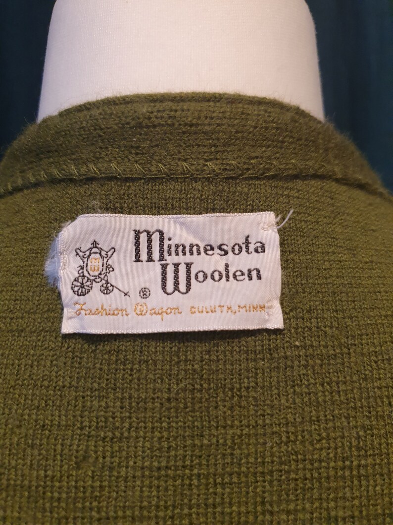 Op de afbeelding: Een groene wollen trui met een wit label waarop staat "Minnesota Woolen Fashion Wagon Duluth, Minn."