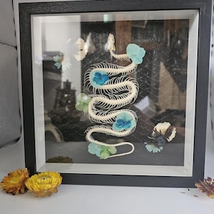 May include: A framed artwork showcasing a snake skeleton, decorated with blue and green flowers. The snake skeleton is centrally placed, with the flowers providing colour against a dark backdrop. The frame is black.
