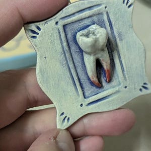 Puede incluir: Colgante de cerámica con un molar detallado dentro de un marco cuadrado. El marco es azul claro con detalles en azul oscuro y un pequeño lazo de metal para colgar. Las raíces del diente están teñidas de rojo.