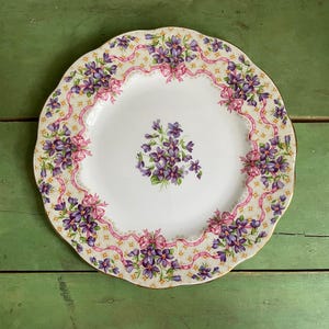 May include: A white ceramic plate with a scalloped edge, adorned with a floral pattern of purple violets and pink ribbons. The plate has a gold trim and a central bouquet of violets.