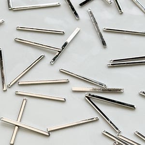 Może przedstawiać: Kolekcja małych, srebrnych metalowych prętów rozrzuconych na białej, prostokątnej powierzchni. Pręty są cienkie i prostokątne, z małym otworem na każdym końcu. Prawdopodobnie są to elementy biżuterii lub materiały rzemieślnicze.