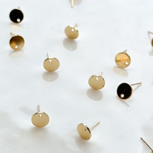 May include: A white plate with a collection of gold-toned earring posts. The posts are round with a small hole in the centre.