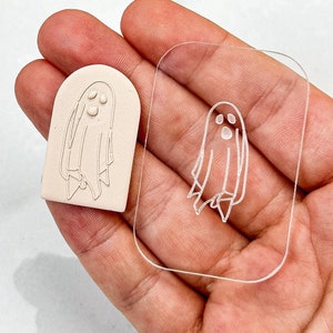 Op de afbeelding: Een witte klei-geeststempel met een transparante acrylstencil. De geest heeft drie ogen en een eenvoudig ontwerp.