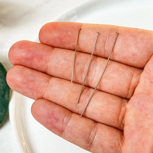 Puede incluir: Cuatro alfileres plateados se sostienen en una mano, con las puntas de los alfileres apuntando hacia arriba.