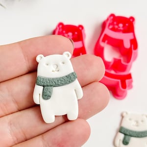 Könnte beinhalten: Eine Sammlung von Bären-Ornamenten. Ein weißer Bär mit grünem Schal wird in einer Hand gehalten. Zwei rote Bären-Ornamente und ein weißer Bär mit grünem Schal befinden sich auf einer weißen Oberfläche. Ein Tannenzweig ist im Hintergrund.