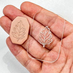 Puede incluir: Un sello de acrílico transparente con un diseño de hoja. El sello se sostiene en una mano, y un sello de goma marrón con el mismo diseño de hoja es visible en el fondo.