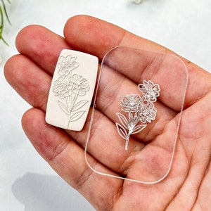 May include: A white polymer clay stamp with a floral design and a clear acrylic stamp with the same design.