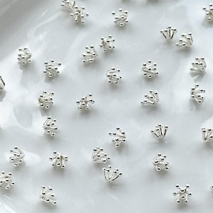 Może przedstawiać: Biały ceramiczny talerz wypełniony srebrnymi metalowymi nasadkami do koralików kwiatowych. Nasadki mają gwiazdkowy wzór z wieloma punktami.