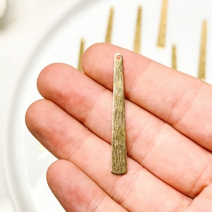 Puede incluir: Primer plano de un colgante alargado de color latón con una superficie texturizada. El colgante tiene un pequeño agujero en la parte superior para ensartar. Varios colgantes similares están dispuestos en un plato blanco, lo que sugiere suministros o componentes para la fabricación de joyas.