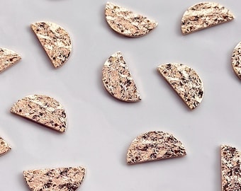 May include: A collection of gold-coloured, half-moon shaped charms with a textured finish. The charms are scattered on a white surface, with floral sprigs in the background. These small metal charms are suitable for jewellery making or craft projects.
