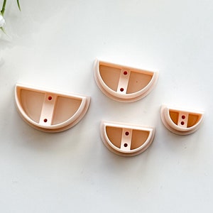 Könnte beinhalten: Vier rosafarbene, halbrunde Keksausstecher aus Kunststoff in verschiedenen Größen. Die Ausstecher haben zwei kleine Löcher in der Mitte.