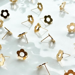 Peut inclure: Une collection de fermoirs de boucles d'oreilles en forme de fleur dorés avec un petit trou au centre de chaque fleur. Les fermoirs sont disposés sur une assiette blanche.